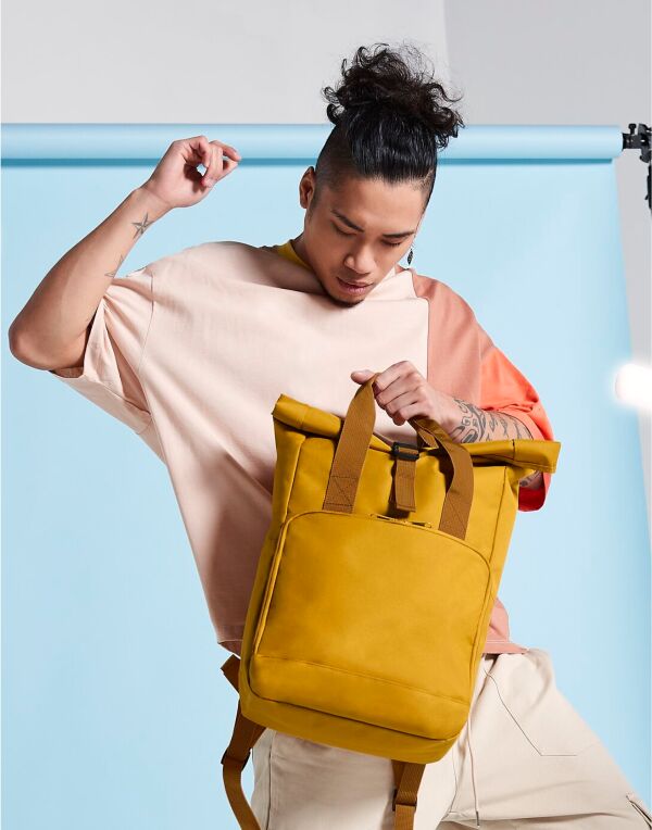 A person carrying the black Bagbase Twin-Handle Roll-Top Backpack by its top handles.