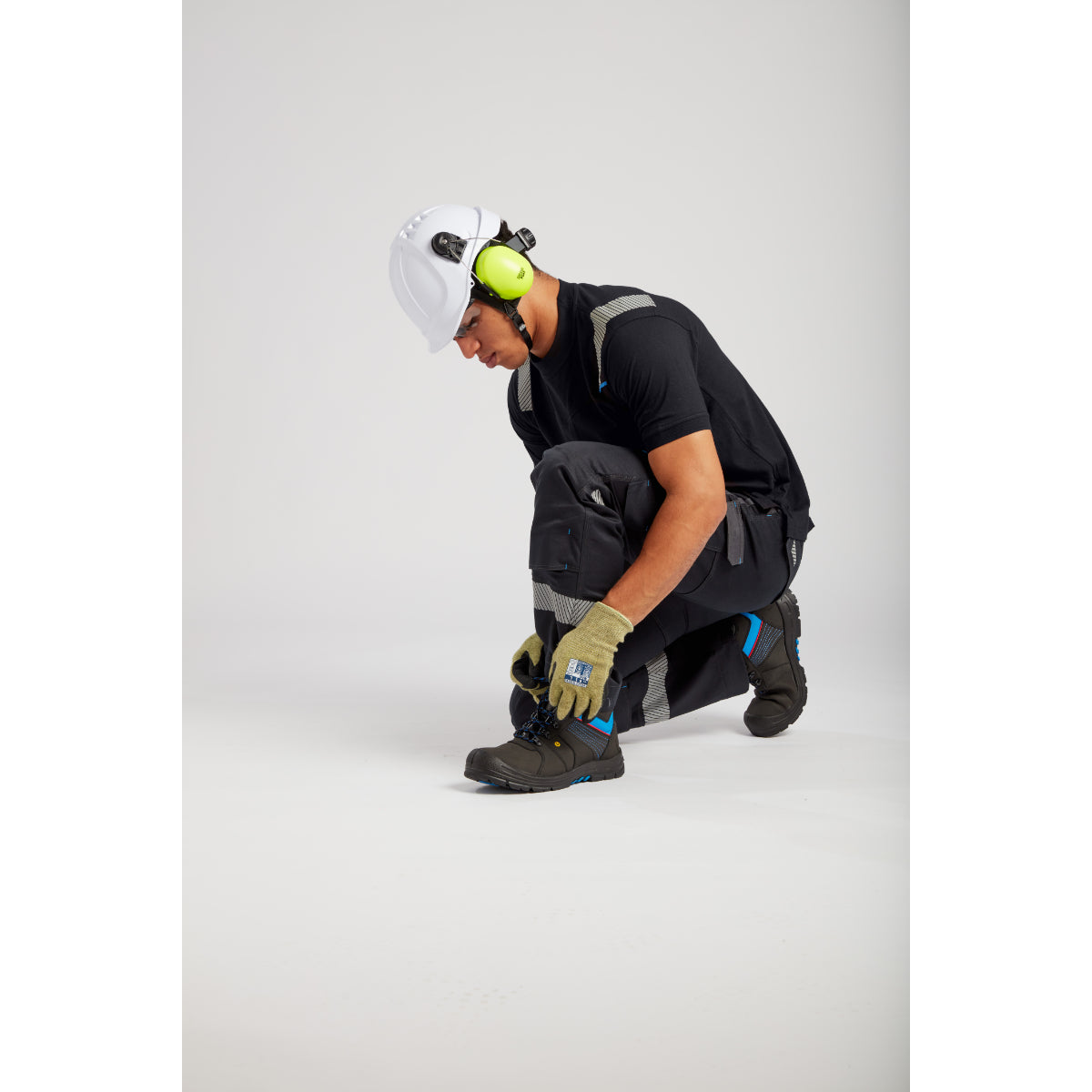 Kneeling worker in safety gear adjusting footwear, wearing helmet with ear protection and reflective workwear