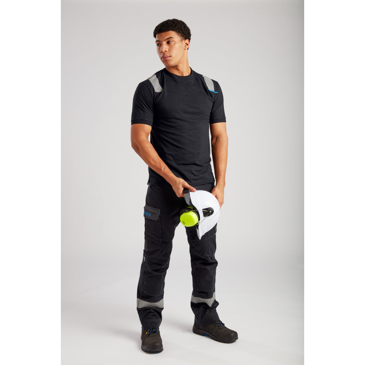 Man holding white safety helmet with green ear protection, wearing black tactical shirt and cargo pants