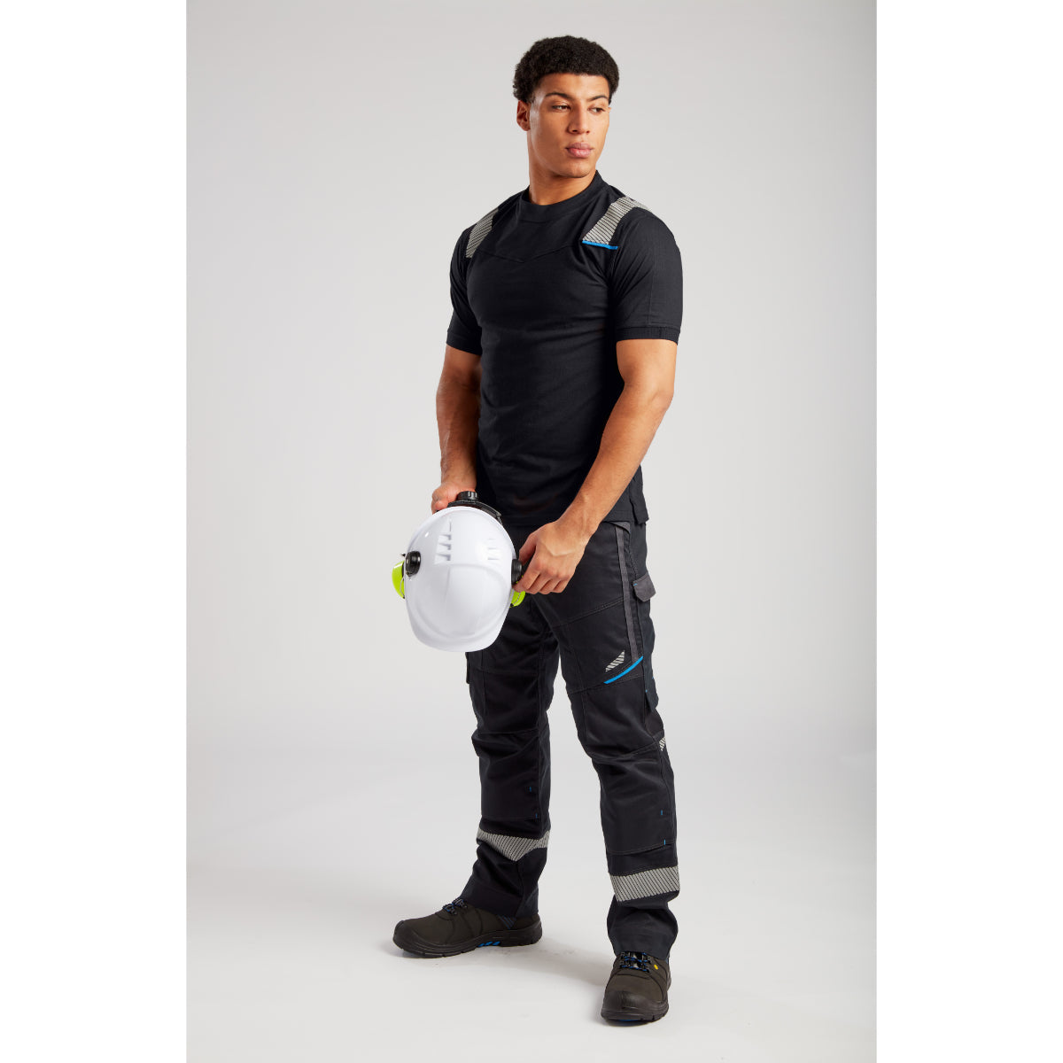 Construction worker with safety harness holding white helmet, wearing black T-shirt and cargo work pants
