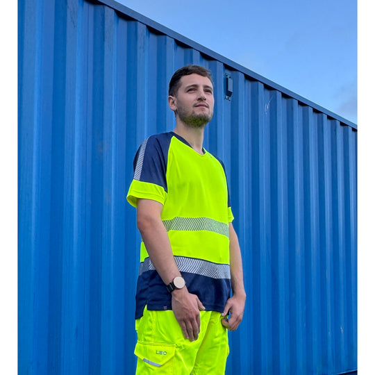Man wearing Leo Workwear Forda EcoViz Performance+ Dual Colour T-Shirt in high-visibility yellow and navy, standing in front of a blue shipping container.