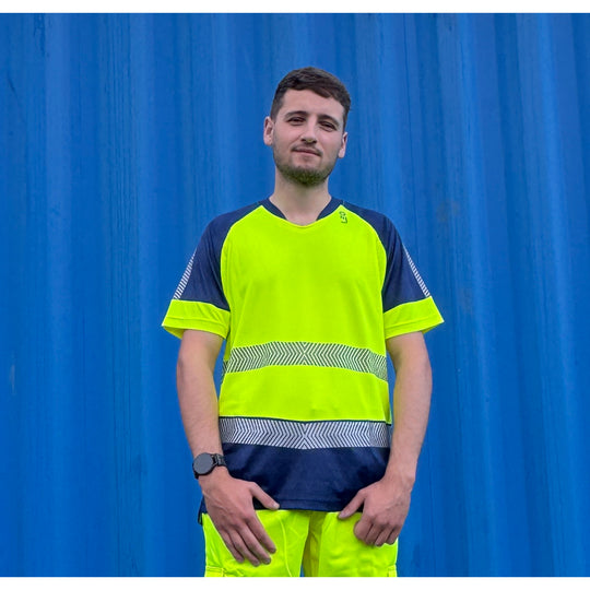 Man wearing Leo Workwear Forda EcoViz Performance+ Dual Colour T-Shirt in high-visibility yellow and navy, standing in front of a blue shipping container.